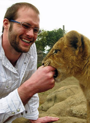 Zach smiles at a young lion, who it gently mouthing his hand.