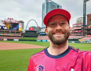 Zach in his bright uniform on poses on the playing field, with the Arch behind him.